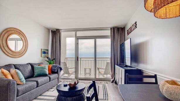 A modern living room with a grey sofa, a round coffee table, a flat-screen TV, and a balcony with a view.
