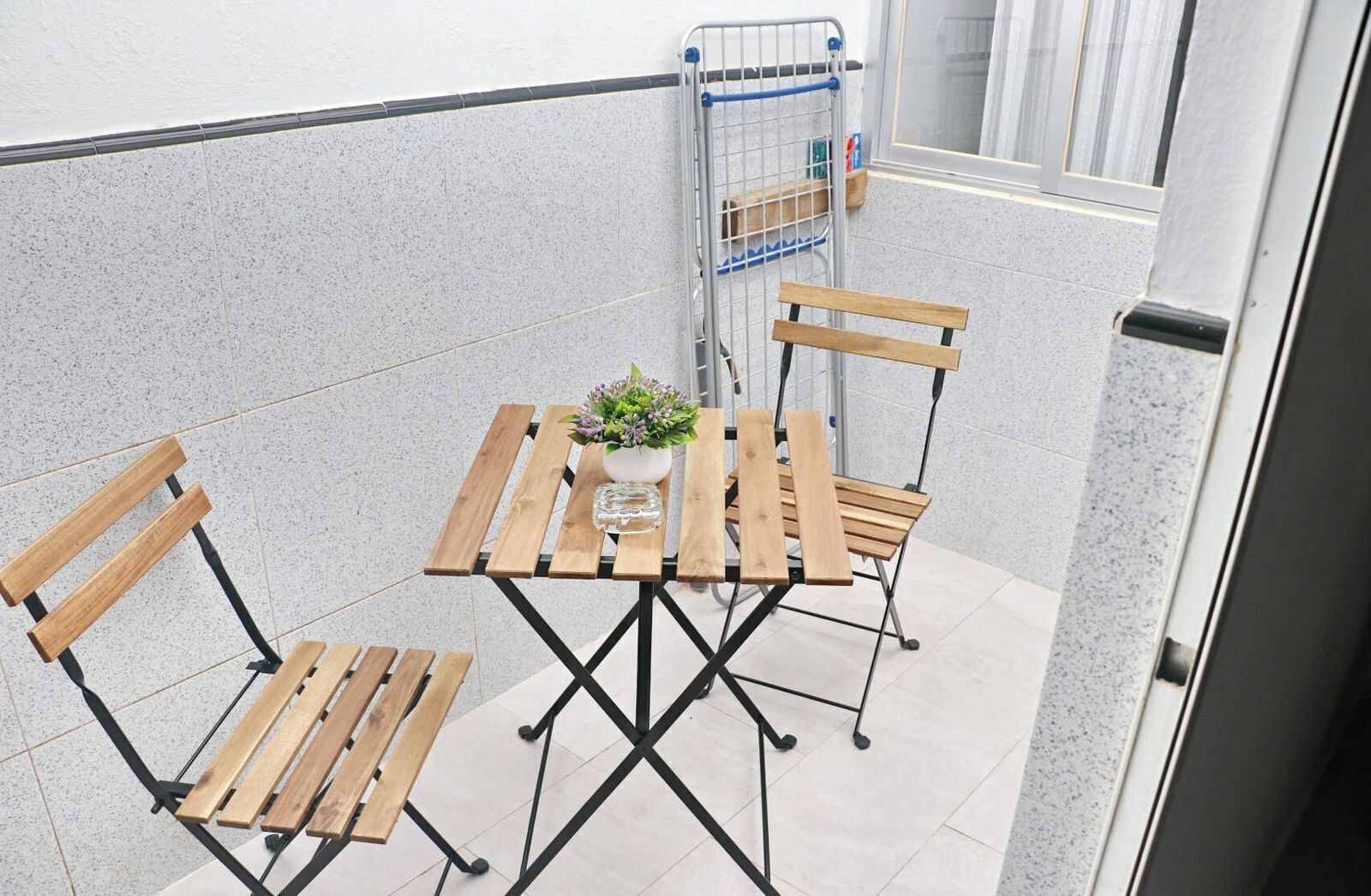 Ein kleiner Sitzbereich im Freien mit einem Holztisch und Stühlen, einem Blumentopf und einem Metallregal mit Gegenständen darauf.