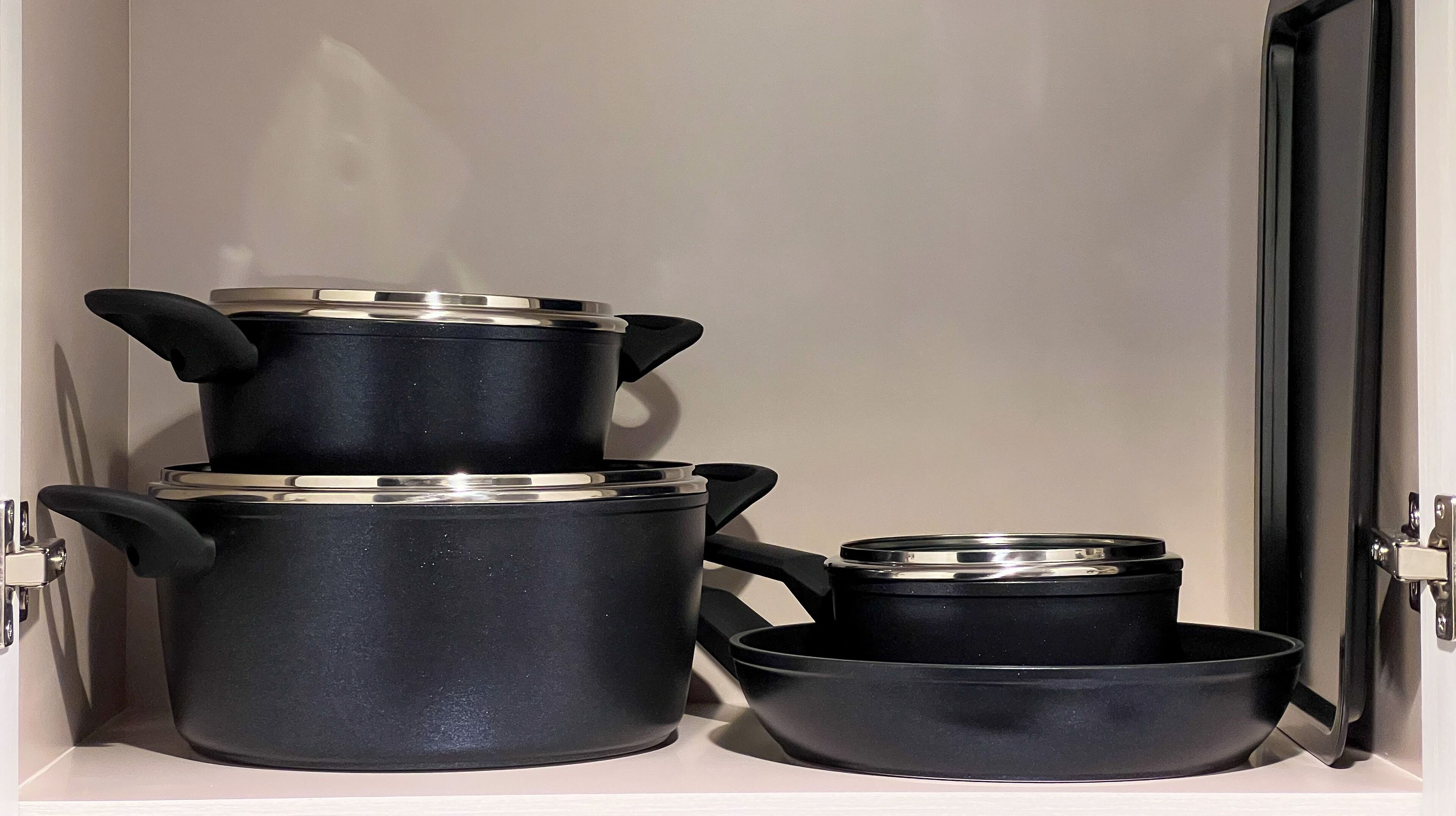 A kitchen cabinet with three black pots and pans, each with a lid.
