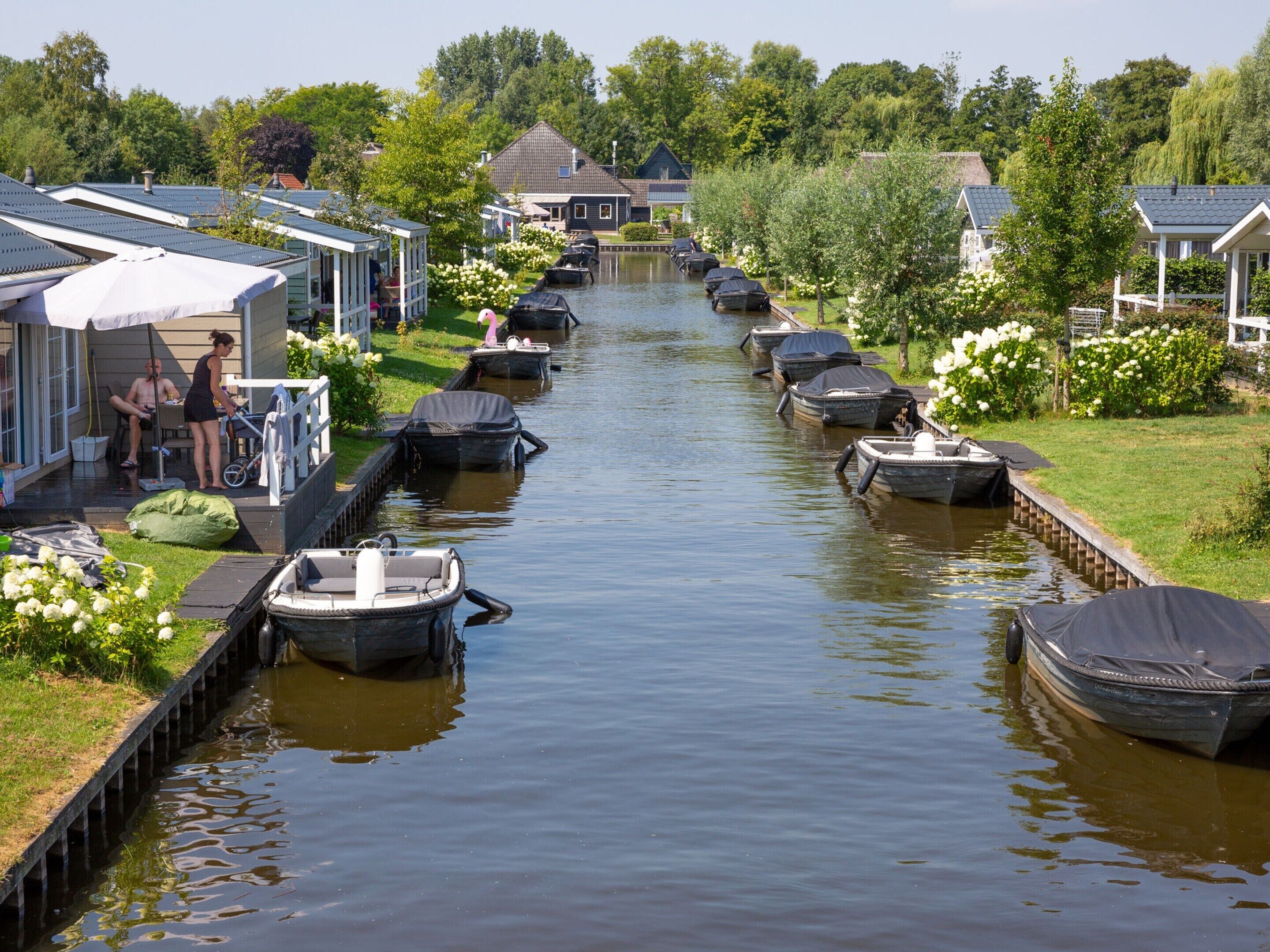 A canal with boats docked on both sides, surrounded by houses with sloped roofs and a grassy area with flowers.