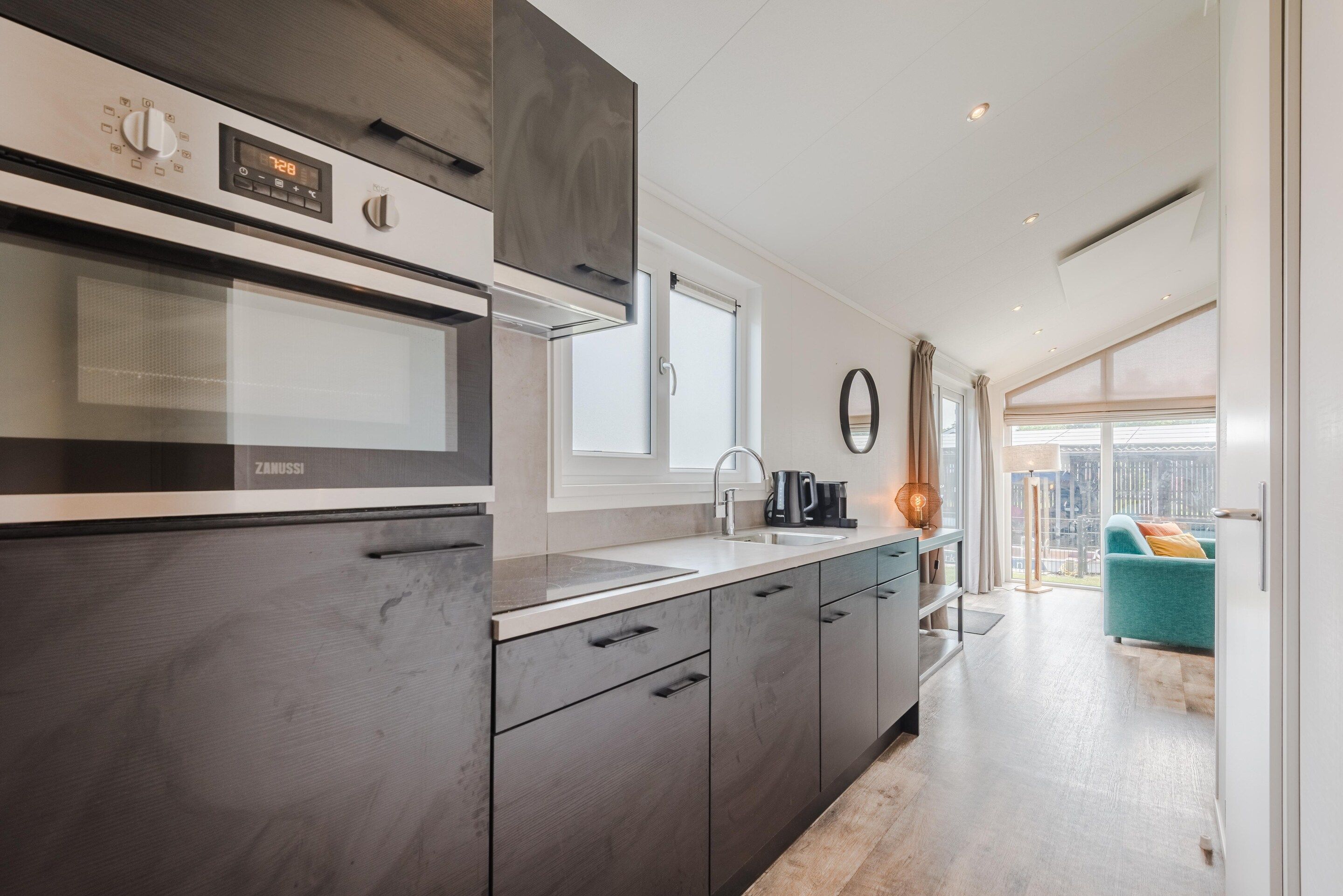 Eine moderne Küche mit Einbauofen, Mikrowelle und Spüle. Über der Spüle befindet sich ein Fenster und man hat einen Blick auf den Balkon mit einer Couch.