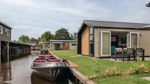 Une maison moderne avec un bateau amarré au bord de l’eau, une pelouse et une terrasse aménagée avec du mobilier d’extérieur.