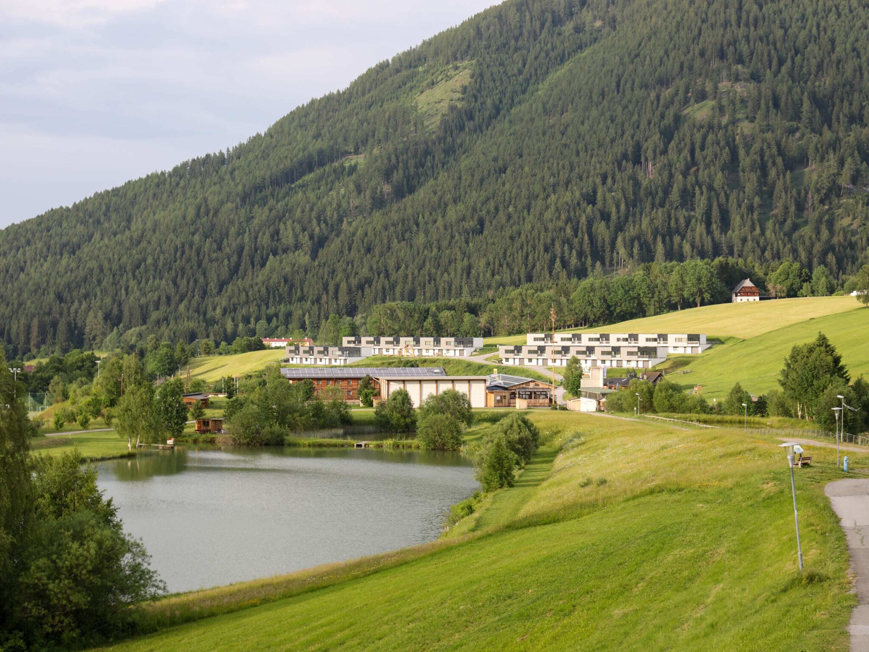 A lakeside hotel with modern buildings, a grassy area, and a forested hill in the background.