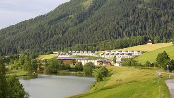 A lakeside hotel with modern buildings, a grassy area, and a forested hill in the background.