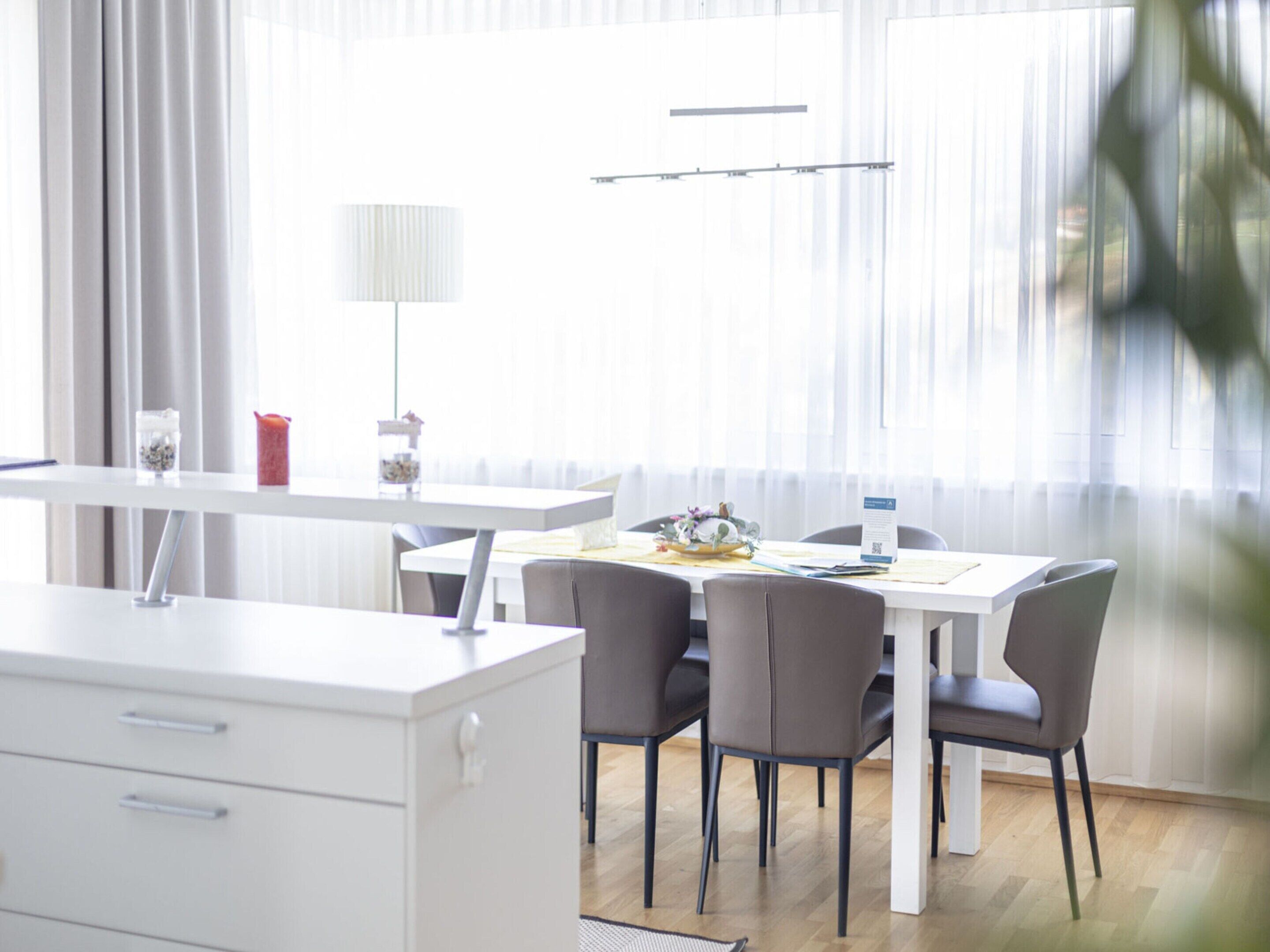 A modern dining area with a white table, grey chairs, a white cabinet, and a window with sheer curtains.