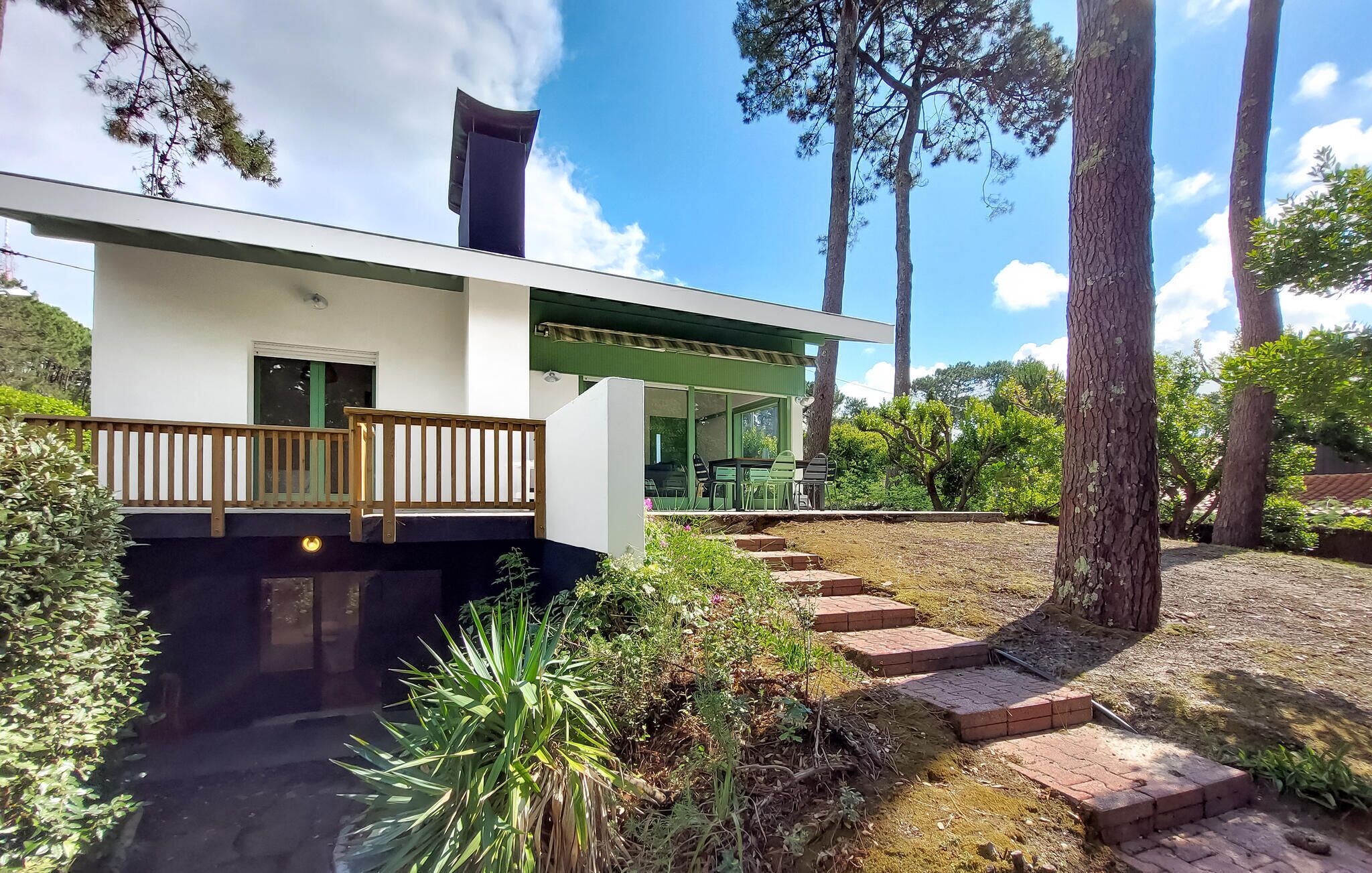 A modern two-story house with a balcony, a covered outdoor seating area, and a garden with steps leading to the entrance.