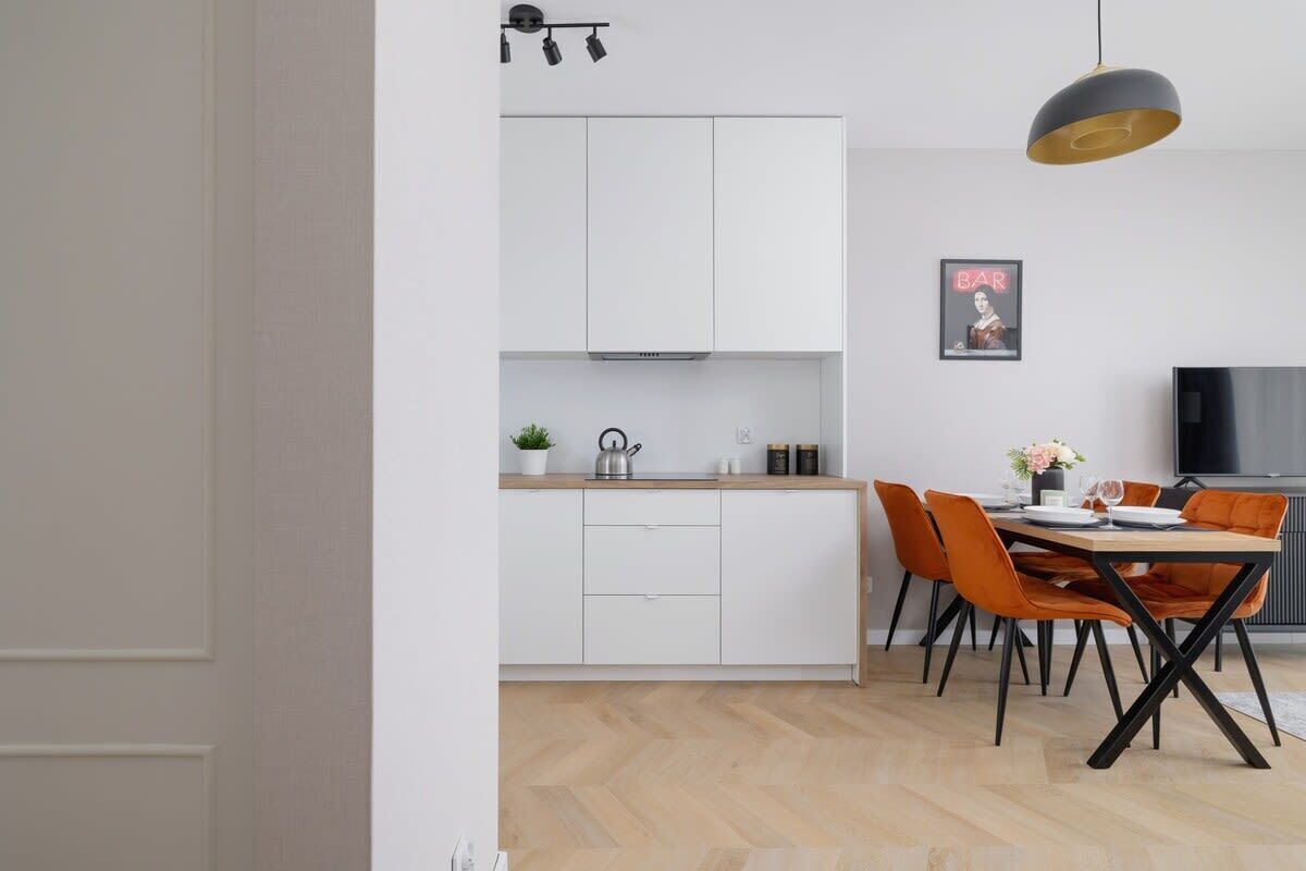 A modern kitchen with white cabinets, a black range hood, and a dining area with orange chairs and a wooden table.