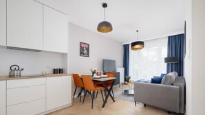 Una cocina moderna con armarios blancos, una mesa de comedor con sillas anaranjadas y un sofá gris en una sala de estar con un ventanal y cortinas azules.