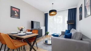Una sala de estar moderna con un sofá gris, sillas naranjas, una mesa de comedor preparada para cuatro personas, un televisor y un amplio ventanal con cortinas.