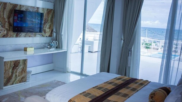 A modern hotel room with a large bed, a flat-screen TV mounted on a marble wall, and a balcony with a view of the ocean. - Stay And Homely Quintessential Hotel (Abuja)
