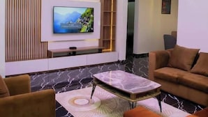 A modern living room with a flat-screen TV, a chandelier, and a sofa. - Stay And Homely Quintessential Hotel (Abuja)