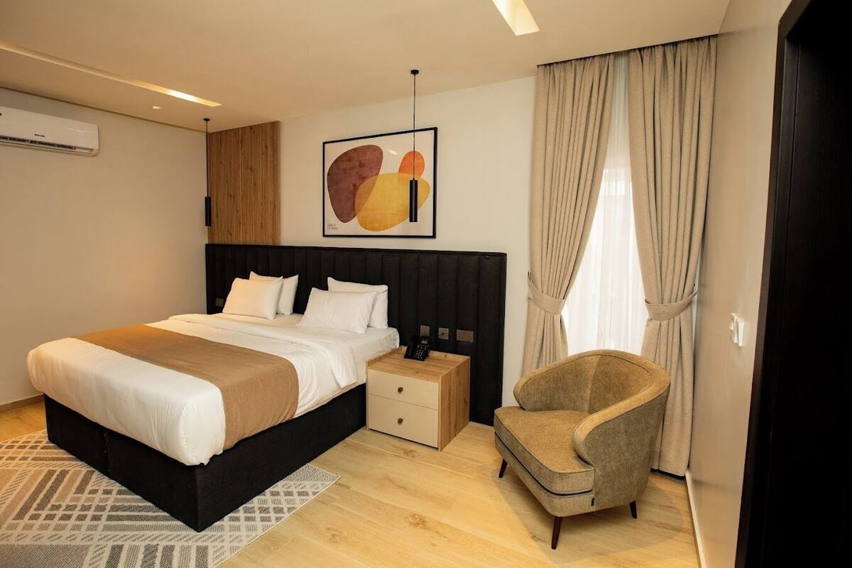 A modern hotel room with a large bed, bedside tables, a chair, and a wall-mounted air conditioner.
