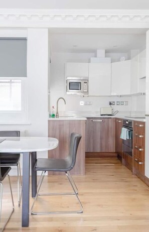 A modern kitchen with white cabinets, a microwave, and a toaster. There is a dining table with chairs and a wooden floor. - Central 2BD Flat, 10 Mins to Buckingham Palace! (London)