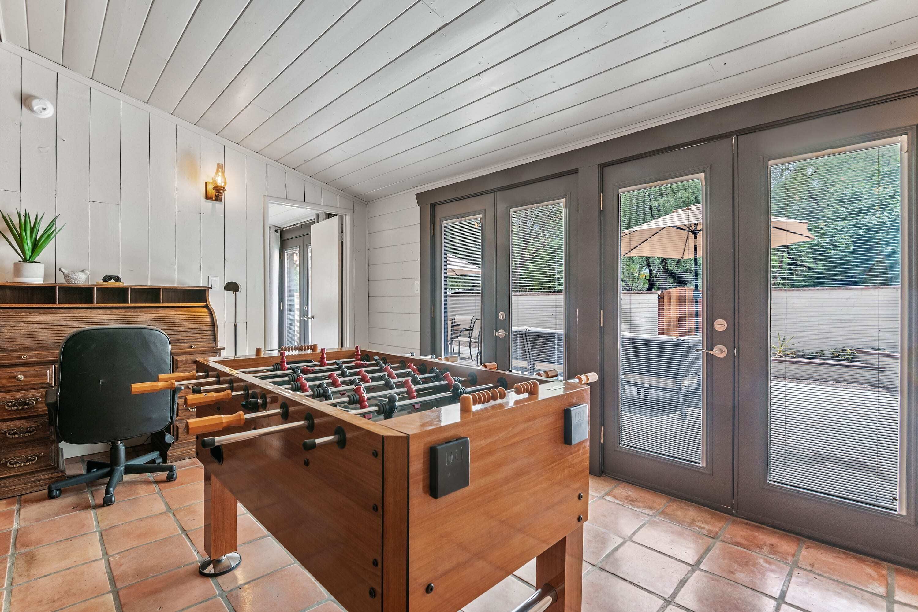 Una habitación con mesa de futbolín de madera, un escritorio con cajones, una silla y una planta en maceta.