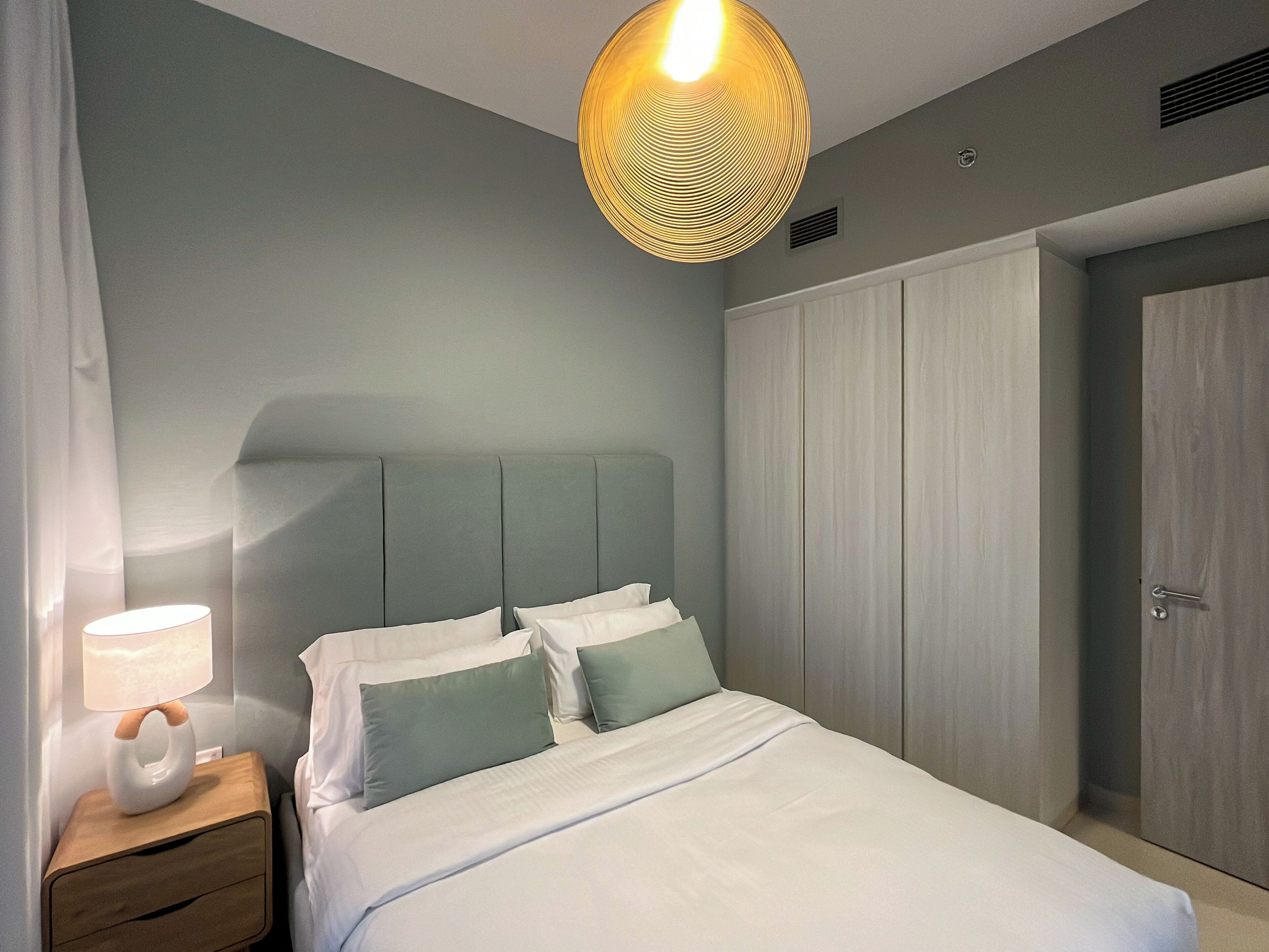 Ein Schlafzimmer mit Bett, Nachttisch, Lampe, Kleiderschrank und einer Pendelleuchte.