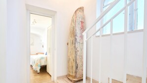 A hallway with a wooden surfboard leaning against the wall, a bed with a blue and white bedspread, and a staircase with white railings. - Casa Alessandra in Anacapri (Anacapri)