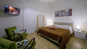 Una camera da letto con un letto, una sedia, una TV, un comodino e uno specchio fissato alla parete.