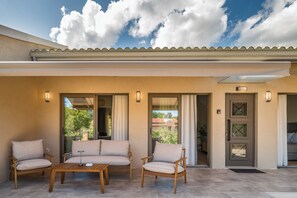 Un espace extérieur abrité avec trois chaises et une table, une porte dotée d’un panneau en verre et une vue dégagée sur le ciel et les nuages.