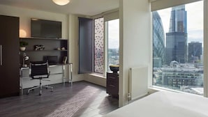 Un dormitorio moderno con una cama grande, un escritorio con una silla, un televisor y vistas a un paisaje urbano a través de un ventanal amplio.
