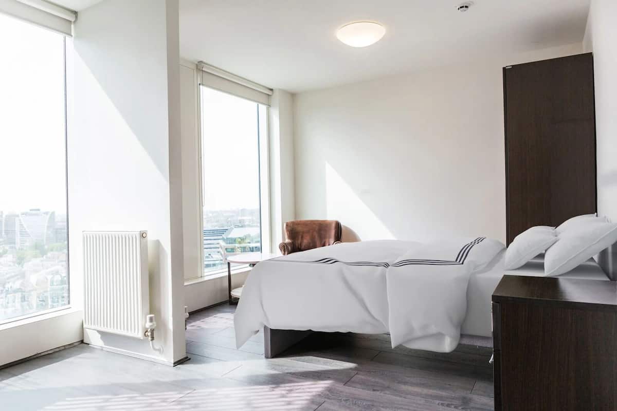 Ein modernes Hotelzimmer mit einem großen Fenster, einem Bett mit weißer Bettwäsche, einem Holzstuhl und einer dunklen Holzkommode.