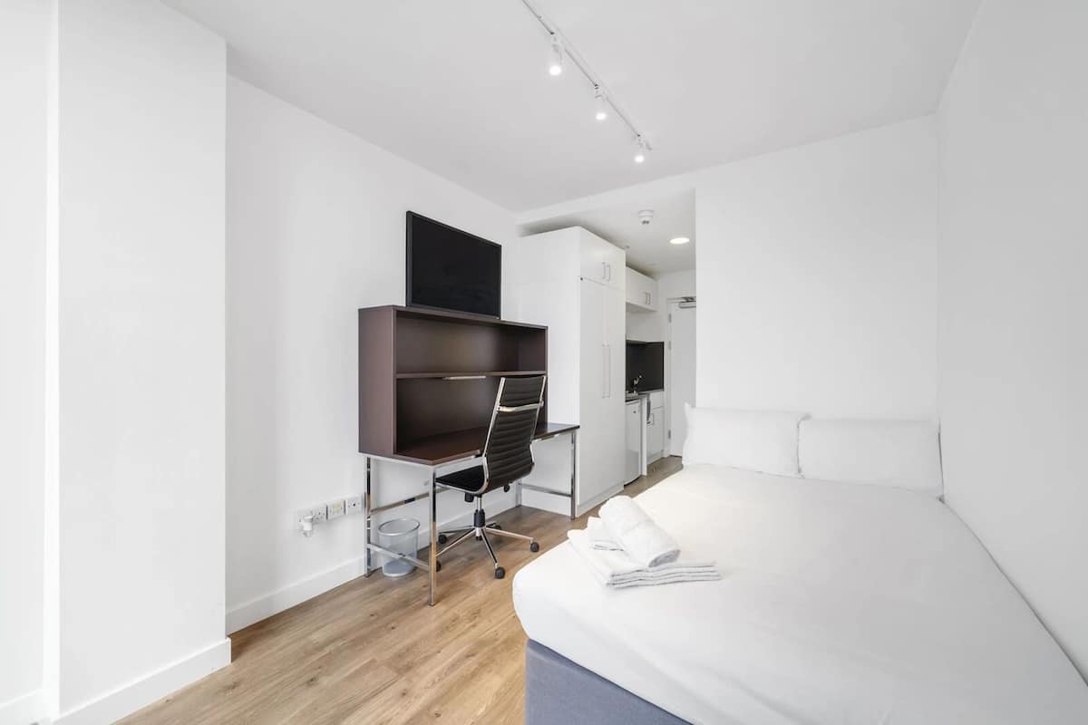 Ein modernes Hotelzimmer mit Bett, Schreibtisch mit Stuhl, Kleiderschrank und einer kleinen Küchenzeile.