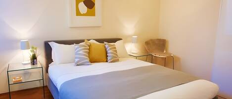 A modern bedroom with a large bed, two bedside tables, a chair, and a wall-mounted artwork.