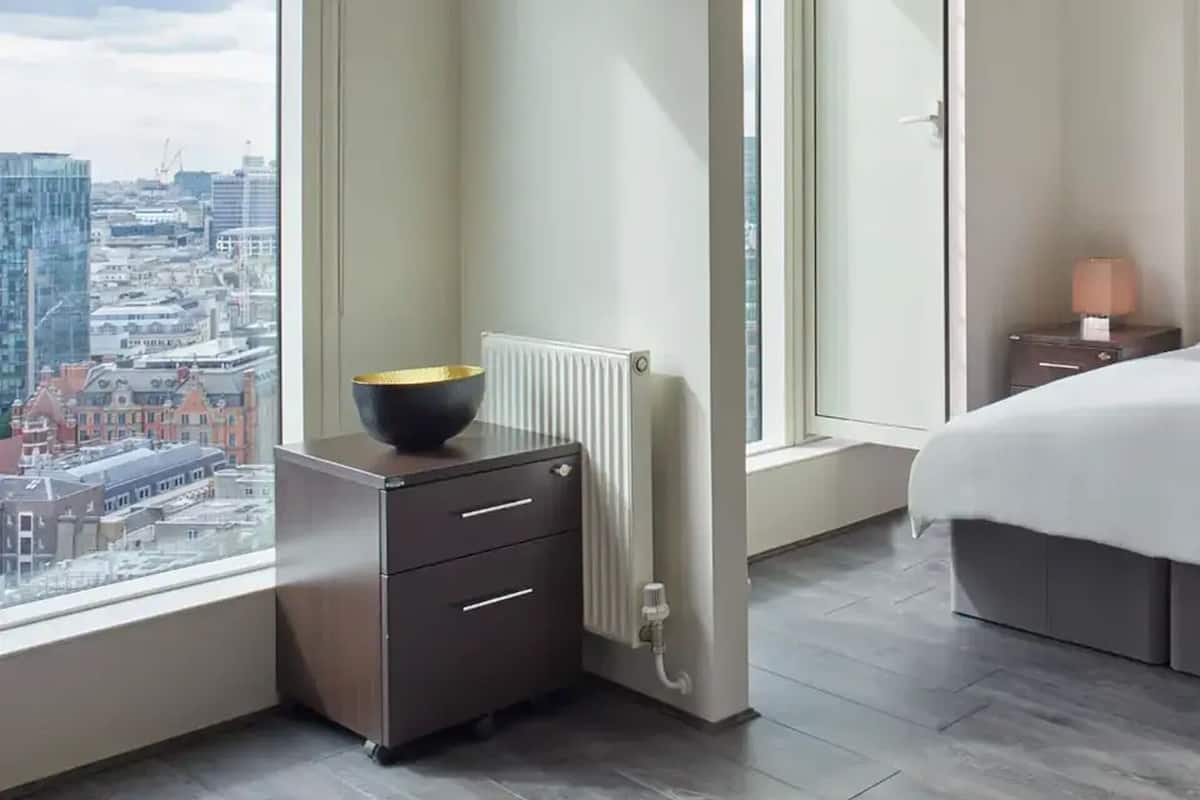 Ein modernes Schlafzimmer mit Blick auf die Stadt, ausgestattet mit Heizkörper, Nachttisch mit Lampe und Bett mit weißer Daunendecke.
