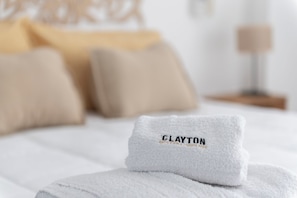 A neatly folded white towel with the word "CLAYTON" embroidered on it, placed on a bed with beige pillows and a white bedspread. - LMC 509 (Buenos Aires)