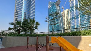 A playground with a slide and climbing structures, set against a backdrop of modern buildings and palm trees. - Chic 1BR Retreat in Business Bay (Dubai)