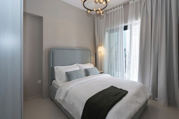 A modern bedroom with a large bed, a nightstand, a lamp, and a view of buildings through sheer curtains.