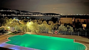 A swimming pool with a green tint, surrounded by trees and a view of a cityscape at night. - Le Terrazze PT36 by Wonderful Italy (Toscolano Maderno)