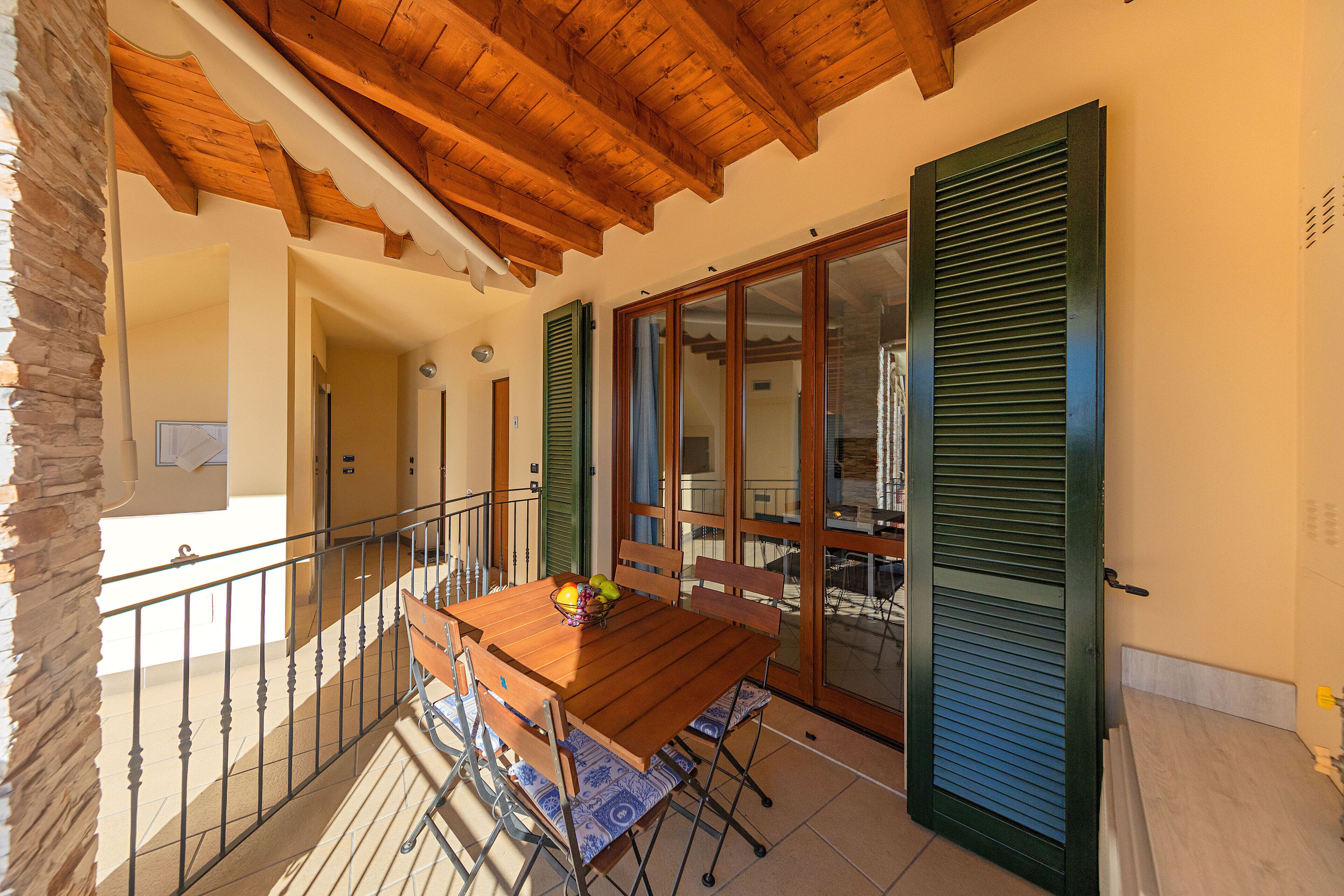 A balcony with a wooden table and chairs, a stone wall, and a sliding glass door with shutters.