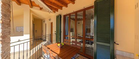 A balcony with a wooden table and chairs, a stone wall, and a sliding glass door with shutters.