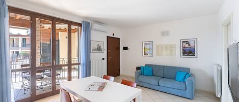 A modern living room with a white dining table, blue sofa, and a large window overlooking a balcony.