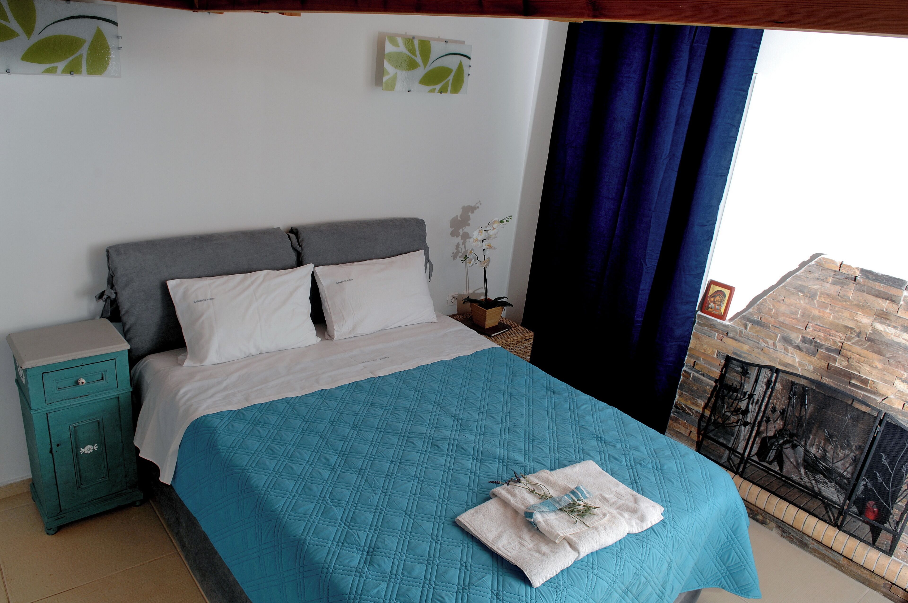 A bedroom with a bed, a nightstand, a window with curtains, and a view of a stone wall.