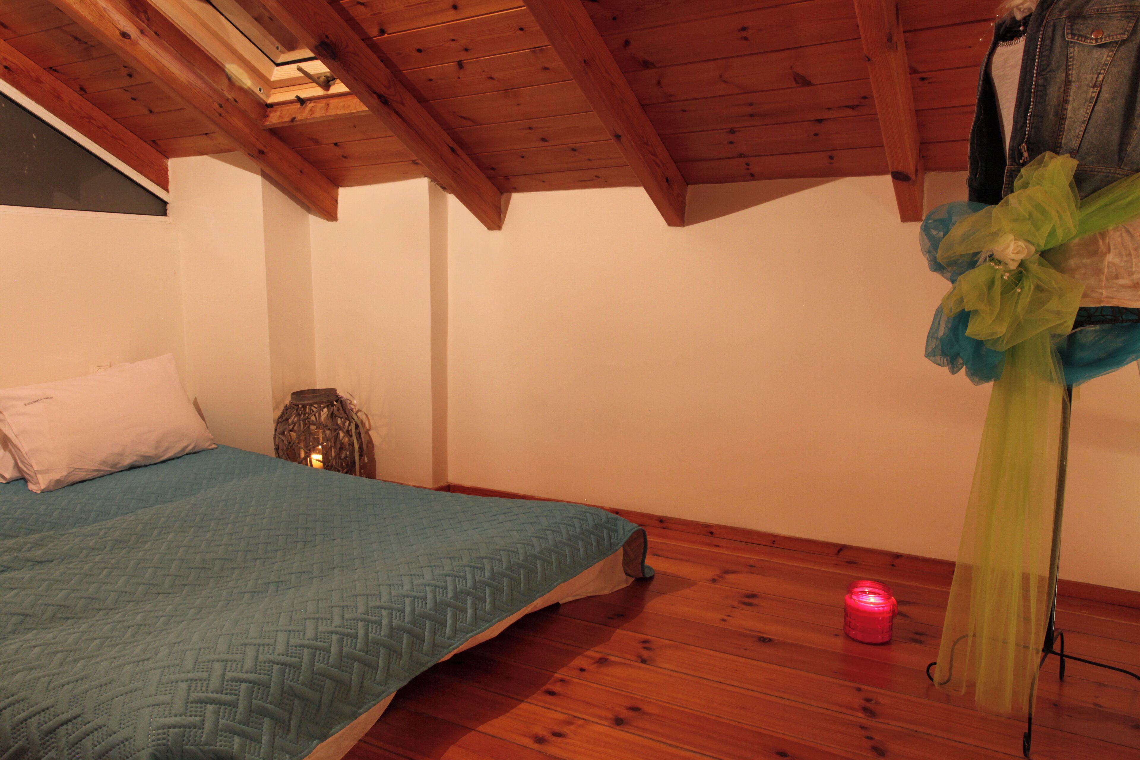 Ein Schlafzimmer mit Holzbalkendecke, einem Bett mit blauer Steudecke, einem kleinen Tisch mit Kerze und einem blau-grünen Dekorationselement an der Wand.