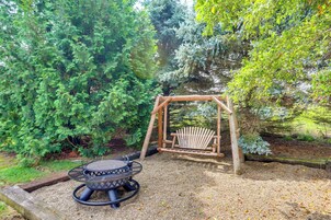 Un jardín con un columpio de madera, una mesa redonda con estructura metálica y un brasero.
