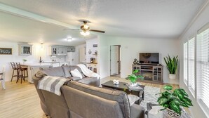 A spacious living room with a sofa, coffee table, and potted plants. A kitchen with white cabinets and a dining area with chairs are visible in the background. - North Fairfield Family Home w/ Hot Tub & Porch! (North Fairfield)