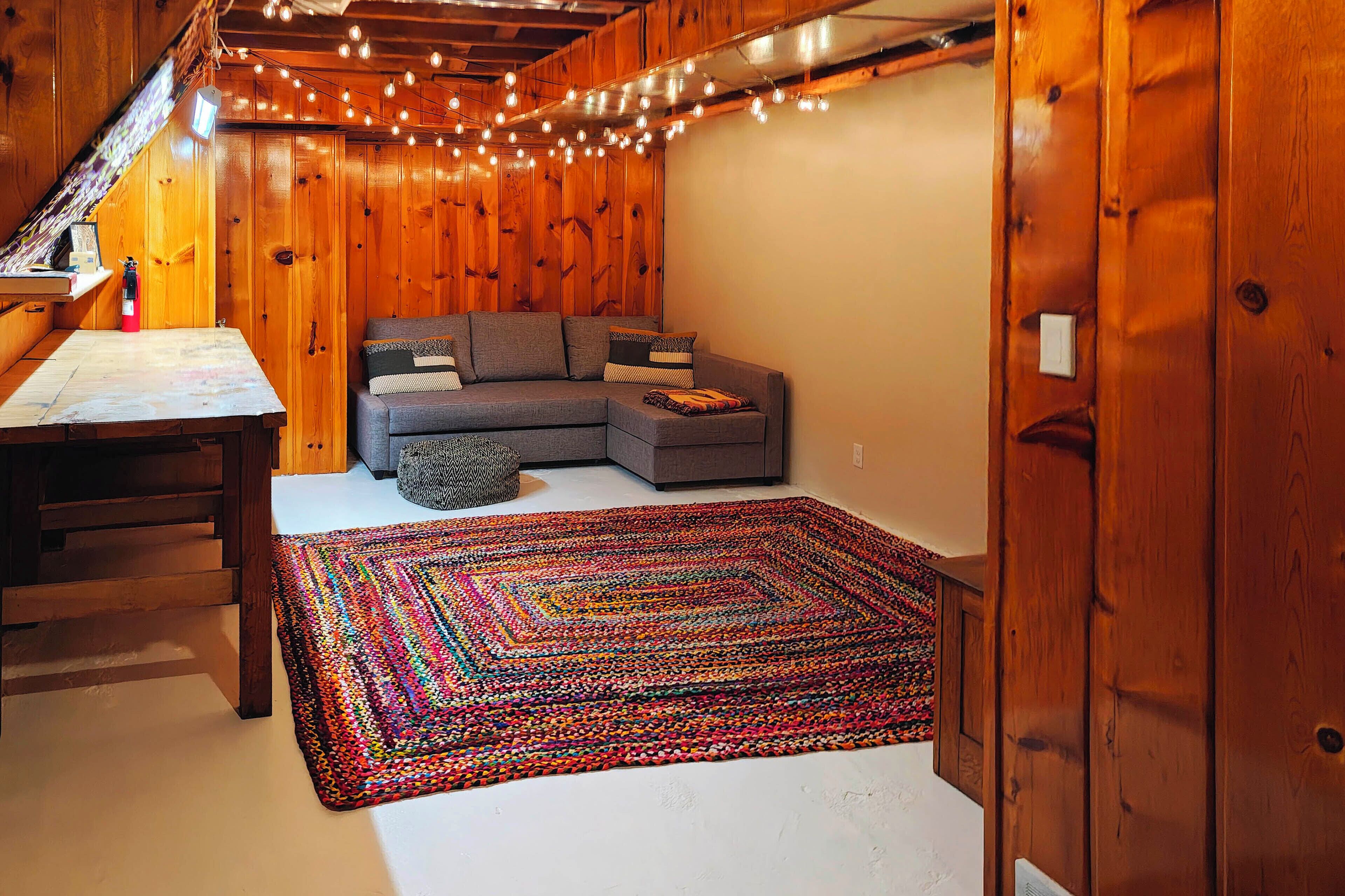 Une pièce chaleureuse avec un plafond en bois, un canapé d’angle gris, un tapis coloré tissé et une table avec une lampe.