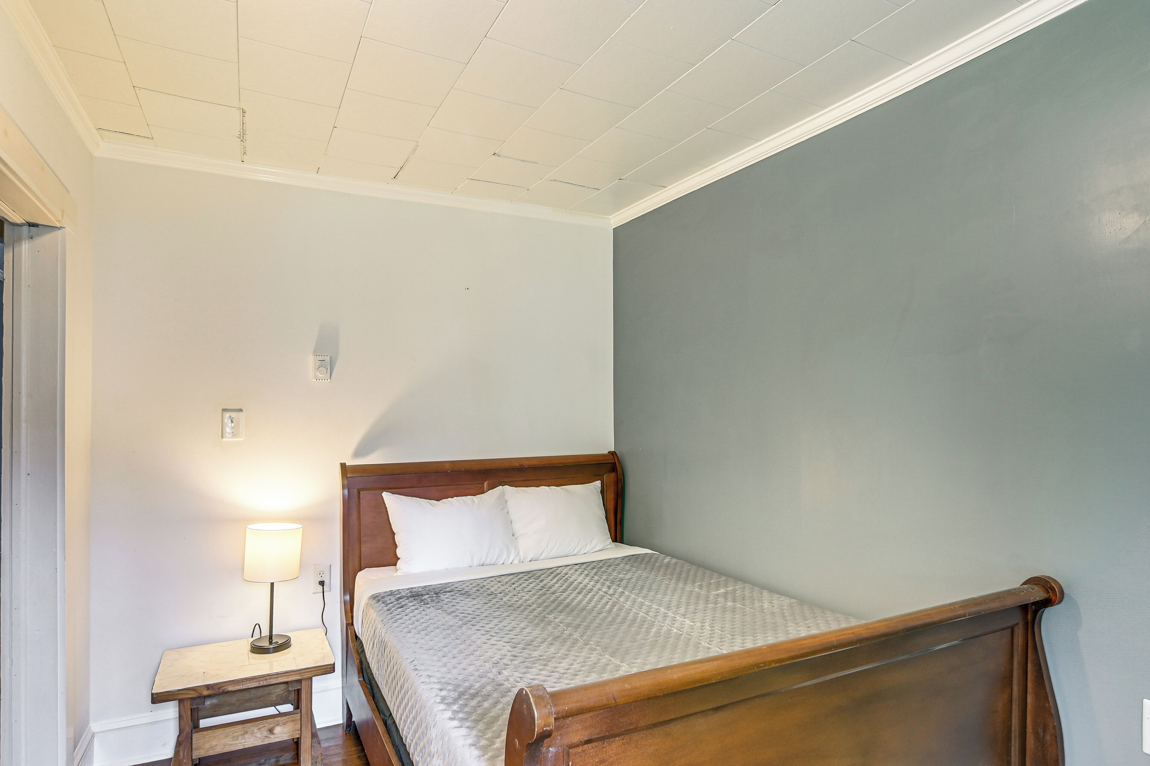 Quarto com cama de madeira, mesa de cabeceira com abajur e parede lisa.