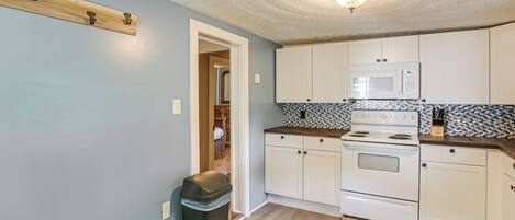 A kitchen with white cabinets, a microwave, and a stove. There is a trash can and a wooden shelf on the wall.