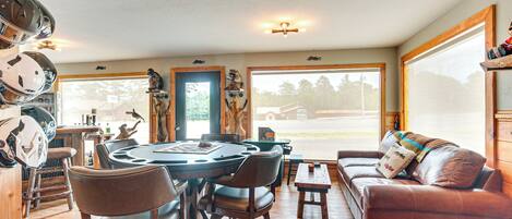 A dining area with a round table, chairs, and a sofa. There are helmets on the wall, a wooden door, and a large window with a view of the outside.