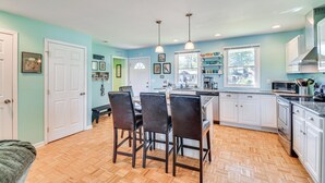 A modern kitchen with white cabinets, a central island with bar stools, pendant lighting, and a view of the outdoors through a window. - Charming Wolcott Cottage w/ Fire Pit & Mini Dock (Wolcott)