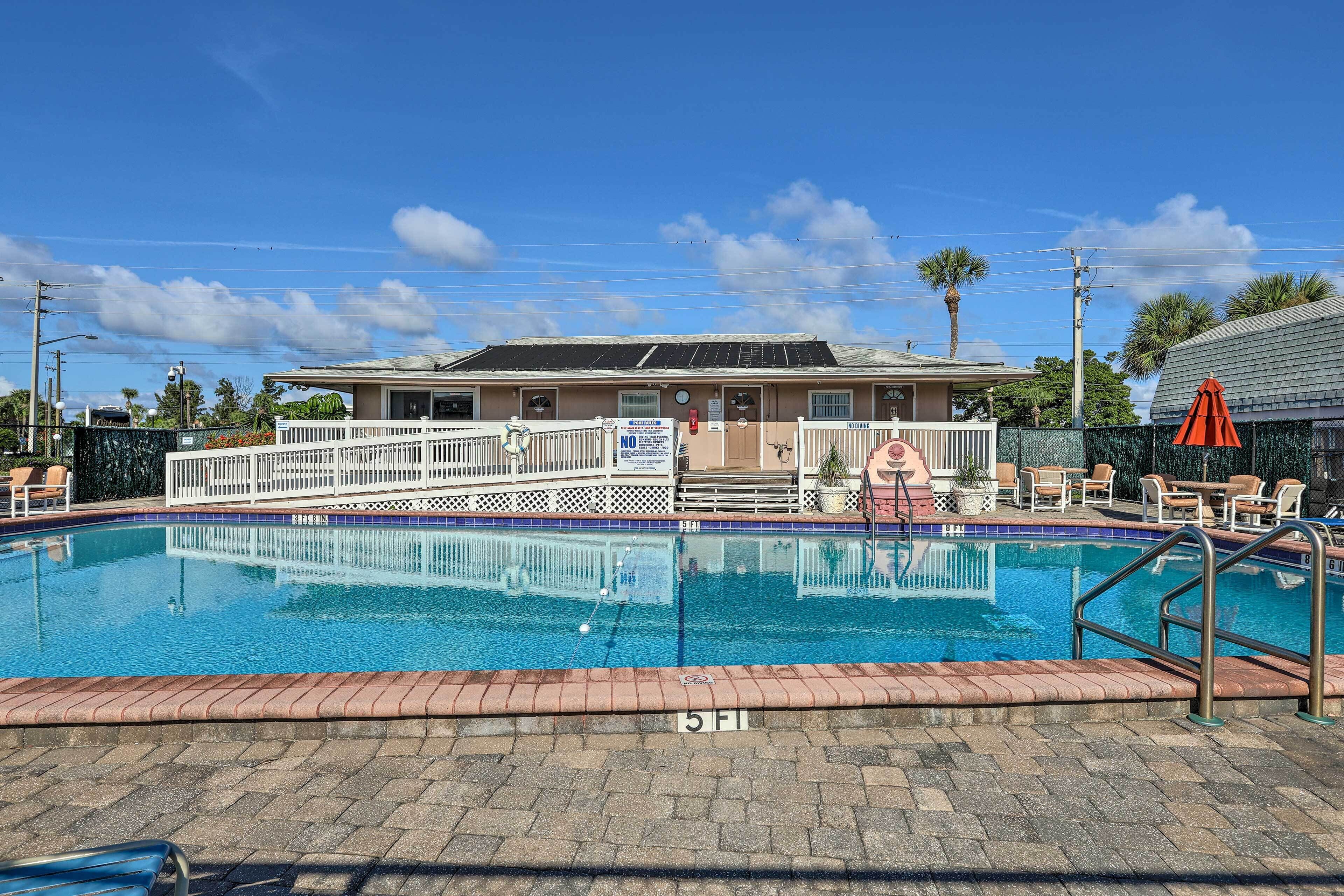 En swimmingpool med et hvidt hegn, liggestole og en bygning med en overdækket terrasse i baggrunden.