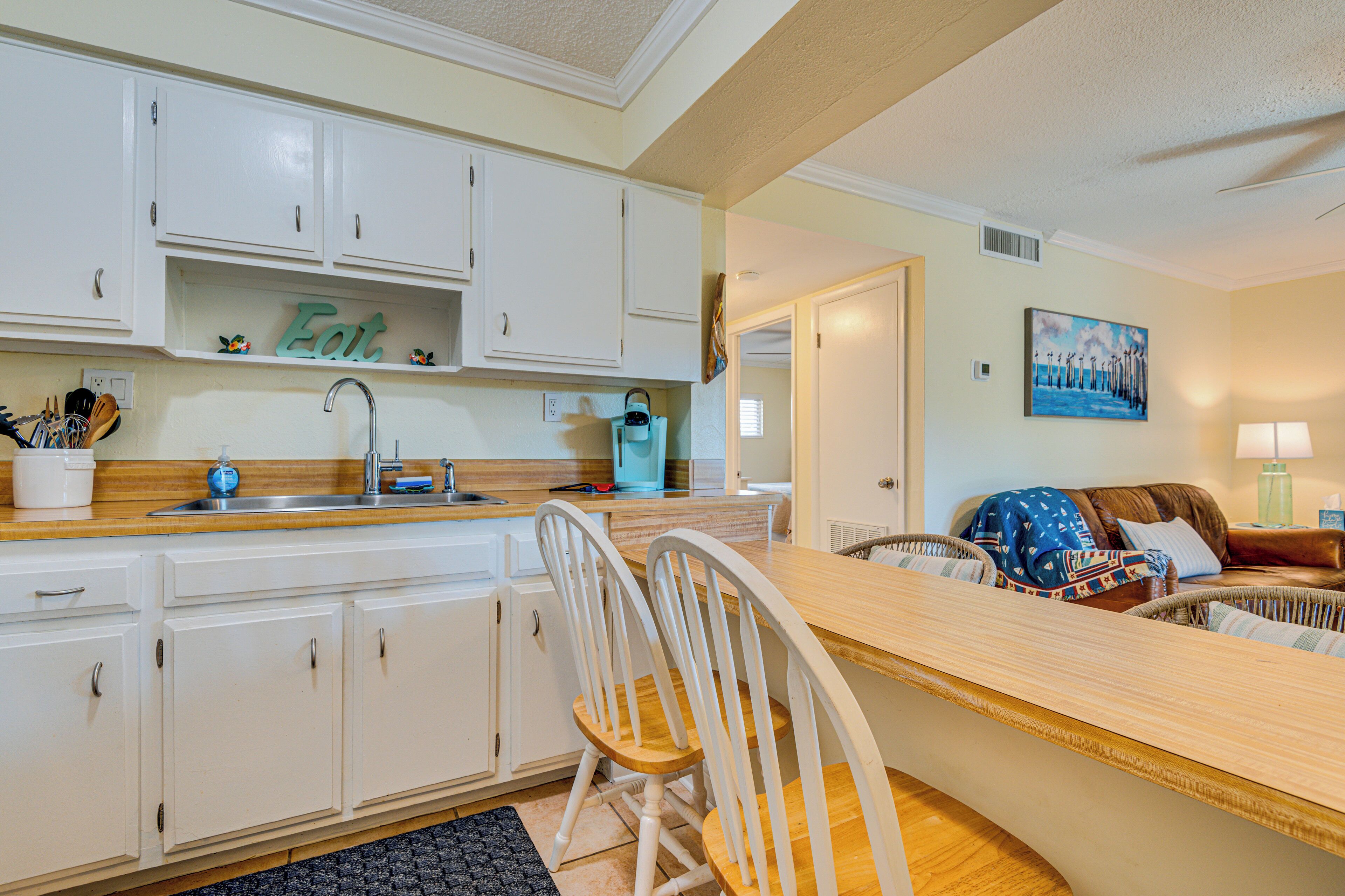 Cocina con armarios blancos, encimera de madera y un cartel que dice 'Eat'. Hay una zona de comedor con una silla de mimbre y un sofá.