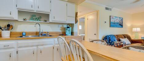 Cocina con armarios blancos, encimera de madera y un cartel que dice 'Eat'. Hay una zona de comedor con una silla de mimbre y un sofá.