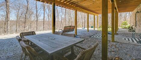 Une terrasse extérieure couverte, avec une table en bois et des chaises en osier, entourée d’une zone recouverte de gravier et offrant une vue sur les arbres.