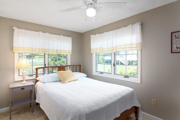 A bedroom with a bed, nightstand, lamp, and two windows with curtains. - Sweet Family Cottage on Beautiful Potter's Pond in East Matunuck (South Kingstown)