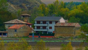 A rural village with traditional stone buildings, a central two-story house with a balcony, and surrounding fields.