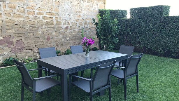A rectangular outdoor table with six chairs, set against a stone wall and surrounded by well-maintained grass and hedges.